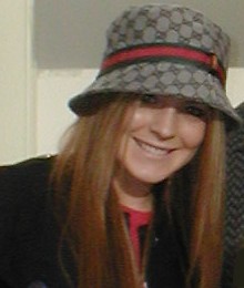 A red-haired Caucasian female wearing a black coat and a hat.